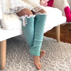 May include: A pair of light blue knitted leg warmers with white lace trim and pearl-like buttons. The leg warmers are worn by a child sitting on a white couch.