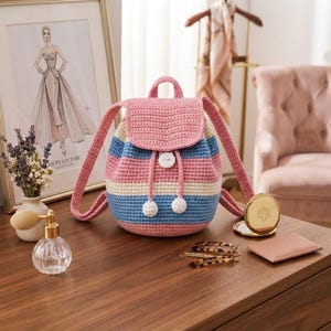 Könnte beinhalten: Ein gehäkelter Rucksack mit horizontalen Streifen in Rosa, Hellblau und Creme. Die Tasche hat einen Klappenverschluss mit einem weißen Knopf, zwei passende Schultergurte und zwei weiße Bommel-Akzente an den Kordelzügen.