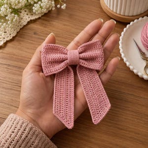 Könnte beinhalten: Ein altrosa gehäkeltes Schleifen-Haaraccessoire mit einem zentralen Knoten und zwei langen, rechteckigen herabhängenden Enden. Die Schleife ist mit einem feinen, gleichmäßigen Maschenbild gefertigt, das eine strukturierte Oberfläche erzeugt.