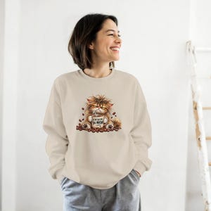 Puede incluir: Una sudadera de cuello redondo en color beige con un gráfico de un gato de pelo largo, despeinado y gruñón, que sostiene una taza de café con el texto JE NE SUIS PAS DU MATIN, rodeado de granos de café tostados sobre un fondo liso.