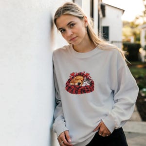 Puede incluir: Una sudadera gris claro de manga larga con un estampado gráfico de un cachorro dorado y un gatito atigrado durmiendo juntos bajo una manta de cuadros rojos y negros, decorada con pequeños corazones rojos flotando sobre los animales.