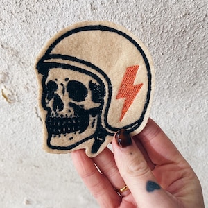 May include: A tan felt patch featuring a black skull wearing a motorcycle helmet. A red lightning bolt is on the helmet. The patch is held up by a hand.