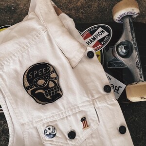 May include: Off-white denim vest featuring a black skull patch with "SPEED" text and a skull pin. The vest rests on a skateboard with a "Champion Park Plugs" sticker and a skateboard wheel.