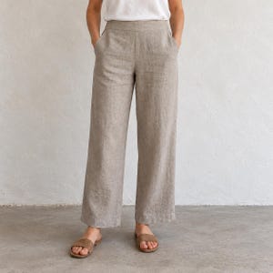 May include: High-waisted wide-leg trousers in a neutral beige tone, featuring a clean waistband, side pockets, and a straight-cut silhouette that falls to the ankle. The garment is paired with a simple white t-shirt and brown sandals.