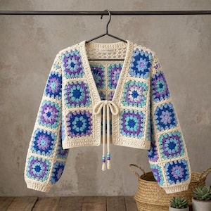 May include: A cropped cardigan made of cream-colored yarn featuring a granny square pattern in shades of blue, purple, and turquoise. The garment has long sleeves, a V-neckline, and a front tie closure with two small beads at the ends.