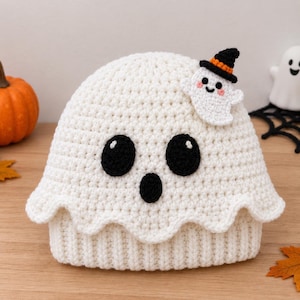 May include: A white crocheted beanie featuring a ghost face with two large black circular eyes and a smaller black circular mouth. A small white ghost applique wearing a black and orange witch hat is attached to the upper right side.