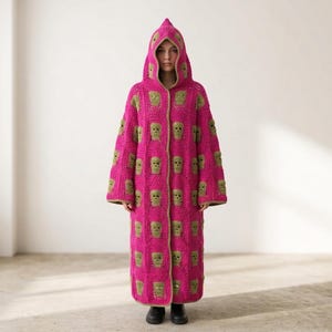 Könnte beinhalten: Ein langer, gehäkelter Kapuzen-Cardigan in leuchtendem Fuchsia-Pink mit einem sich wiederholenden Muster aus hellgrünen Totenkopf-Motiven. Das Kleidungsstück hat lange Ärmel, eine spitze Kapuze und einen geraden Schnitt bis zur Wadenmitte.
