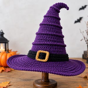 May include: A purple crocheted witch hat featuring a tall, pointed crown with horizontal ridges and a wide brim. A black crocheted band encircles the base of the crown, accented with a square, golden-yellow crocheted buckle at the front center.