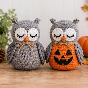 May include: Two crocheted owl figurines made of grey yarn. Both owls have white circular eye patches with closed black eyelashes and orange beaks. One owl is solid grey, while the other has an orange belly with a black felt jack-o'-lantern face.