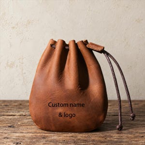 May include: A brown leather drawstring pouch featuring the text Custom name & logo embossed on the front. The pouch has a gathered top secured with a dark brown cord that extends from the side, creating a simple and functional storage accessory.
