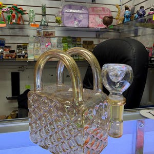 May include: A gold-tinted glass water pipe shaped like a small handbag with a woven texture pattern. The piece features two arched handles on top and a clear glass bowl attachment extending from the side, designed for filtration and smoking use.