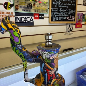 May include: A cylindrical water pipe featuring a vibrant, multi-colored graphic print with comic book-style illustrations. The pipe includes a curved neck, a wide circular base, and a metallic silver-toned nail attachment centered on the top.
