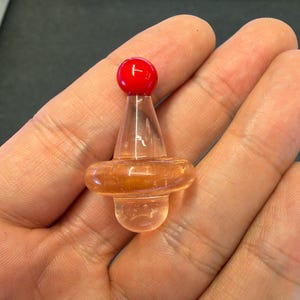May include: A small, clear glass object shaped like a spinning top with a rounded base, a wider middle section, and a bright red spherical bead at the top. The glass has a smooth, polished finish and a transparent quality throughout the structure.