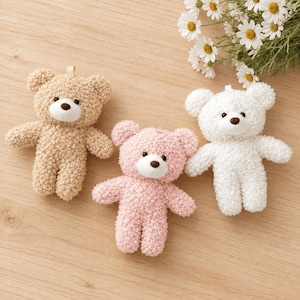 Puede incluir: Tres osos de peluche en beige, rosa claro y blanco, cada uno con un acabado de tela texturizada y rizada, un hocico blanco y una nariz marrón oscuro. Los osos están dispuestos juntos, con un pequeño grupo de margaritas blancas cerca.