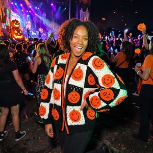 May include: A long-sleeved, button-down cardigan featuring a checkerboard pattern of black and white squares. Each square is adorned with a crocheted orange pumpkin motif with a stitched face. The cardigan has a black ribbed hem and cuffs.