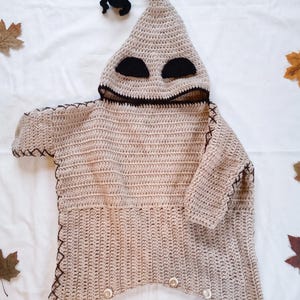May include: A beige crochet hooded garment featuring a pointed hood with a black pom-pom, black oval eye patches, and a wide black mouth opening. The piece includes dark brown cross-stitch detailing along the edges and five light-colored buttons.
