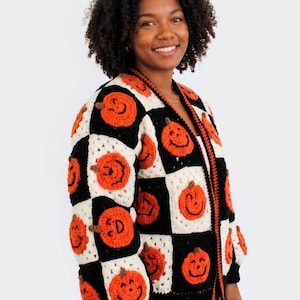 May include: A long-sleeve crochet cardigan featuring a checkered pattern of black and white squares. Each square contains a crocheted orange pumpkin with a smiling face and a brown stem. The cardigan has a black ribbed collar, cuffs, and hem.