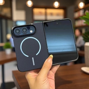 May include: A black foldable smartphone case with a textured back surface, featuring a prominent white circular magnetic charging ring and a rectangular camera module with three lenses and a flash. The case is designed for a folding device.