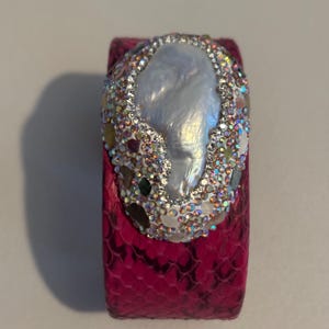 May include: A bright pink cuff bracelet with a textured snakeskin pattern. The centre features a large, irregular white pearl surrounded by a dense cluster of small, iridescent, multi-coloured crystals and various small, smooth, earth-toned stones.