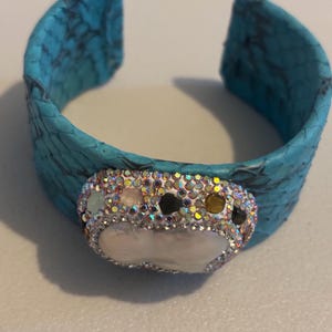 May include: A bright turquoise cuff bracelet featuring a textured, scale-like pattern. The centre is adorned with a large, irregular white pearl surrounded by a border of small, iridescent, multi-coloured crystals and dark stone accents.