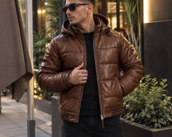 May include: A brown leather puffer jacket featuring a hood with drawstrings, a front zip fastening, and horizontal quilted stitching. The jacket has long sleeves and a high collar, providing a structured fit with a smooth, polished finish.