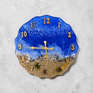 Könnte beinhalten: Eine runde Wanduhr mit gewelltem Rand und einem Harz-Ozean-Design. Die obere Hälfte zeigt tiefblaues Wasser mit weißen Schaumwellen, während die untere Hälfte sandige Strandtexturen mit kleinen Kieselsteinen und zwei Seesternen aufweist. Goldene Ziffern.