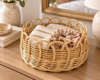 May include: A round, light-colored woven rattan basket featuring a decorative scalloped rim. The basket contains a folded beige fabric item, a tan fabric scrunchie, and a light-colored patterned hair claw clip, all neatly arranged inside.