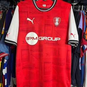 May include: A red soccer jersey with white sleeves, a black and white crew neck, and the text IPM GROUP in a white circle. The shirt features a Puma logo, a team crest, and a repeating RUFC pattern. The number 10 is printed on the inner collar.