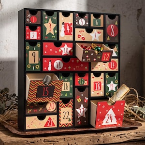May include: A black wooden advent calendar with twenty-four numbered drawers. Each drawer features festive holiday designs including stars, snowmen, and geometric patterns in red, green, and brown. Some drawers are open, revealing small items.