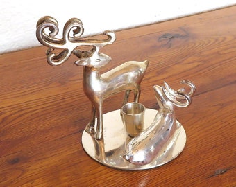May include: A silver-colored metal reindeer candle holder with a small cup for a candle. The reindeer has large, ornate antlers and stands on a round base. Another reindeer figure is on the base. The candle holder is on a wooden surface.