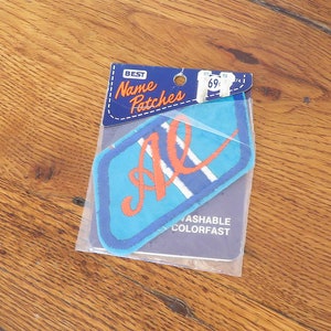 May include: A blue and white iron-on name patch with the letters "AL" in orange. The patch is in a plastic bag with a white label that says "Best Name Patches" and "Washable Colorfast".