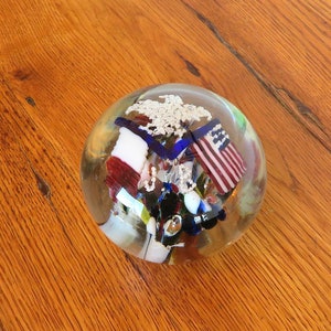 May include: A clear glass paperweight with red, white, and blue glass pieces inside. The glass pieces are arranged to resemble a flag.