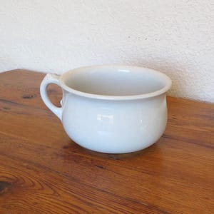 May include: A white, ceramic chamber pot with a single handle. The pot has a rounded shape and a wide opening. The chamber pot is sitting on a wooden surface.