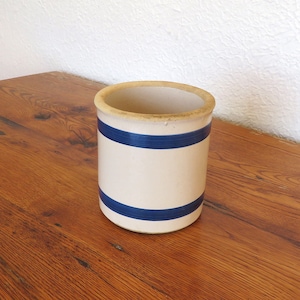 May include: A vintage stoneware crock with a cream-colored base and two navy blue stripes. The rim has a light brown color. This cylindrical container is likely used for food storage or decoration.