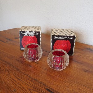 May include: Two clear glass candle holders with a textured surface and a red interior. The candle holders are sitting on a wooden table. The candle holders are packaged in boxes with the text "Snowball Lite" printed on them.