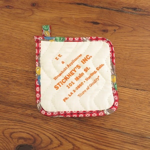May include: A red and white checkered pot holder with a white center featuring the text "I.V. Household Appliances. STICKNEY'S INC. 101 Main St. Ph. LA 2-2660 - Sterling, Colo. Home of Quality" in orange.