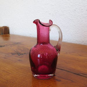 May include: A ruby-red glass pitcher with a clear, curved handle. The pitcher has a ruffled rim and a textured pattern on the neck. The body of the pitcher has a swirling design. The pitcher is sitting on a wooden surface.