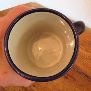May include: A blue-rimmed mug with a white interior, viewed from above. The mug has a dark blue handle and a glossy finish. The mug is sitting on a wooden surface.