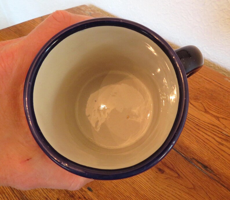 May include: A blue-rimmed mug with a white interior, viewed from above. The mug has a dark blue handle and a glossy finish. The mug is sitting on a wooden surface.