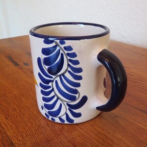May include: A ceramic mug with a white base, adorned with blue leaf-like patterns. The rim and handle are dark blue. The mug sits on a wooden surface. A yellow accent is visible on the side.