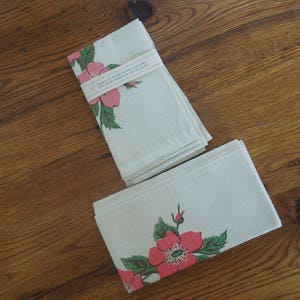 Pu&ograve; includere: Due asciugamani da t&egrave; in cotone bianco con disegni floreali rosa e verdi. Gli asciugamani sono piegati e impilati su una superficie di legno. L'asciugamano superiore ha il testo "Handmade with Love in CA" stampato su di esso.