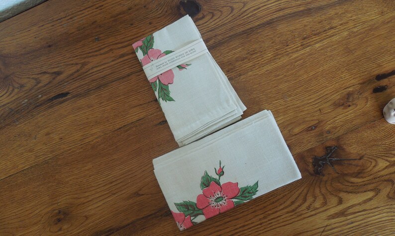 Pu&ograve; includere: Due asciugamani da t&egrave; in cotone bianco con disegni floreali rosa e verdi. Gli asciugamani sono piegati e impilati su una superficie di legno. L'asciugamano superiore ha il testo "Handmade with Love in CA" stampato su di esso.