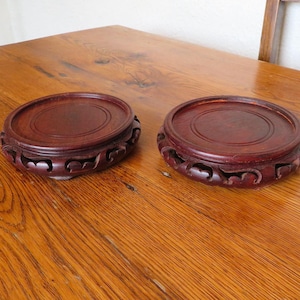 May include: Two round, dark reddish-brown wooden stands with carved floral designs. The stands have a raised center and are displayed on a wooden table. The stands are likely for displaying decorative items.