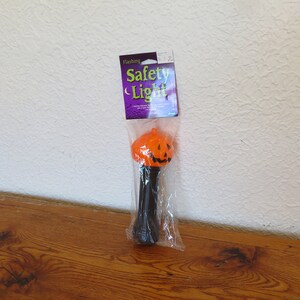 May include: An orange pumpkin-shaped flashing safety light with a black stem, packaged in a clear plastic bag. The pumpkin has a carved jack-o'-lantern face. The product tag reads "Flashing Safety Light".