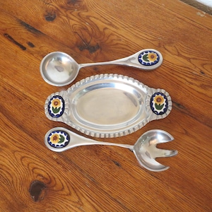 May include: A set of serving utensils and a serving dish made of silver-colored metal. The spoon and fork have decorative ends with a sunflower design. The oval dish has a ridged edge and matching sunflower accents.