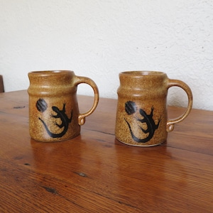 May include: Two brown ceramic mugs with handles. Each mug features a black lizard design with a sun-like circle. The mugs are on a wooden surface.