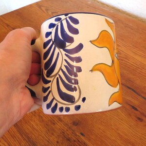 May include: Cream-colored ceramic mug with blue and orange painted designs. The mug features a blue floral pattern on one side and an orange sun-like design on the other. The handle is also blue.