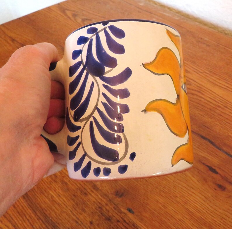 May include: Cream-colored ceramic mug with blue and orange painted designs. The mug features a blue floral pattern on one side and an orange sun-like design on the other. The handle is also blue.