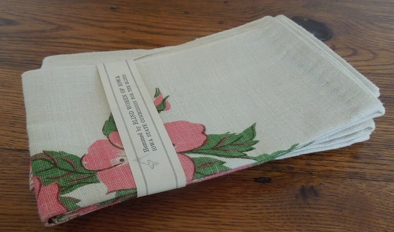 Pu&ograve; includere: Un set di tovaglioli di lino bianco con un motivo floreale rosa e verde. I tovaglioli sono orlati dalle Donne Cieche dell'Iowa, Commissione dello Stato dell'Iowa per i Ciechi.