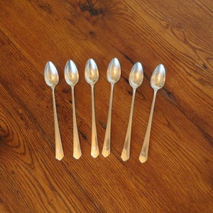 May include: A set of six silver-toned metal spoons arranged in a row. Each spoon features an elongated, slender handle that tapers toward the base and a smooth, oval-shaped bowl. The metal has a polished, reflective finish throughout the design.